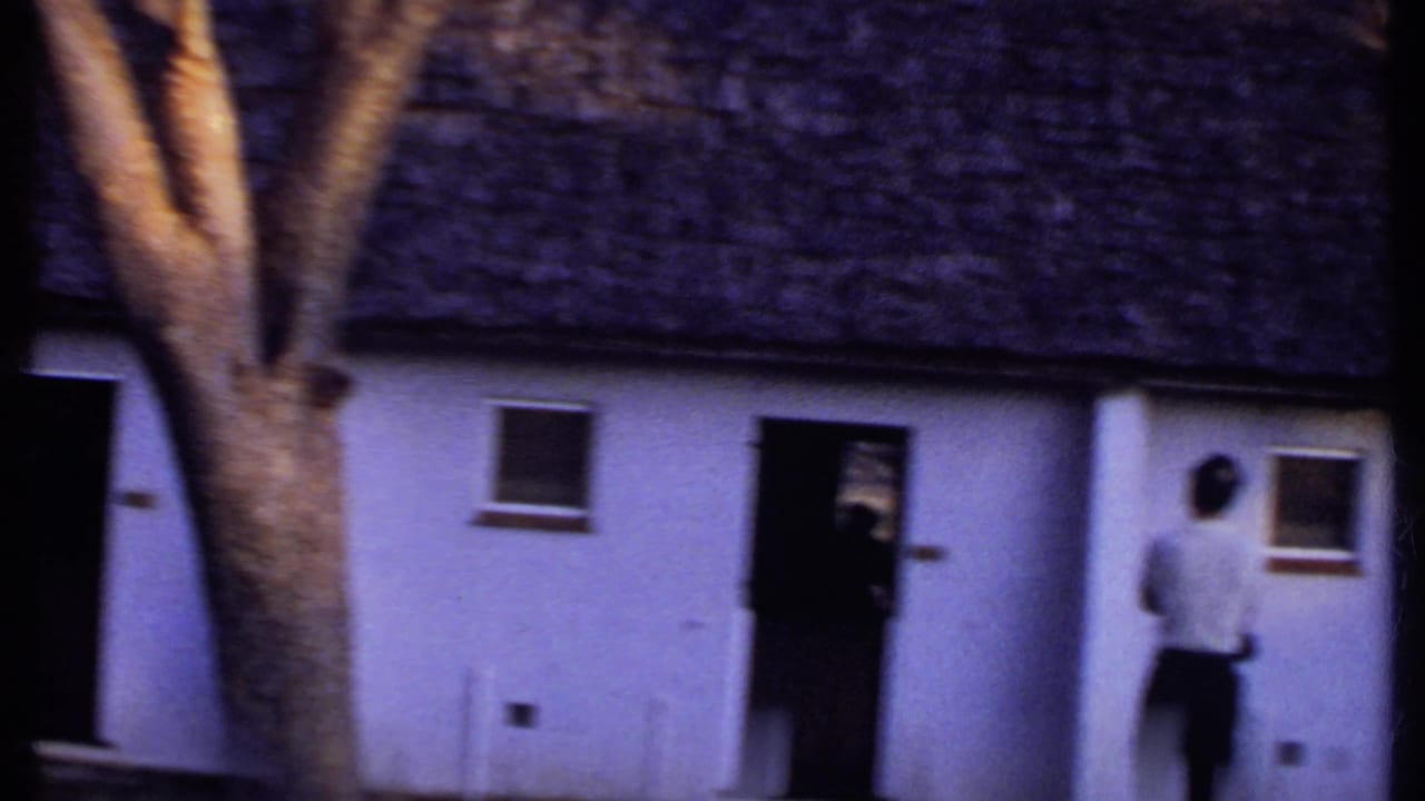 1969 Kenya 8mm frame showing a man at a doorway of a white building with a thatched roof and tree in the foreground.