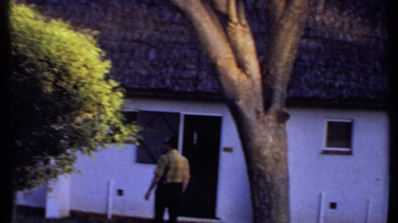 1969 Kenya frame with a man walking toward a doorway on a white thatched-roof building, partly framed by trees and shrubs.