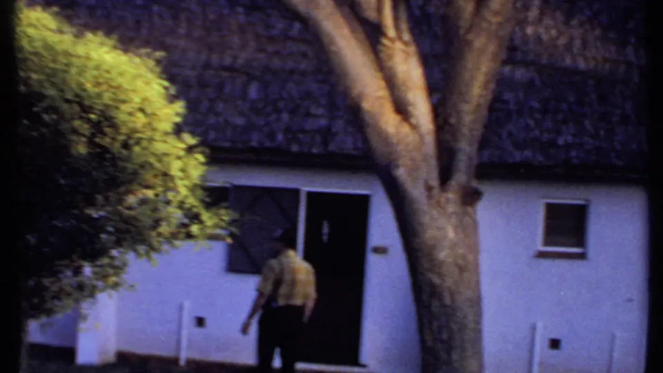 1969 Kenya frame with a man walking toward a doorway on a white thatched-roof building, partly framed by trees and shrubs.