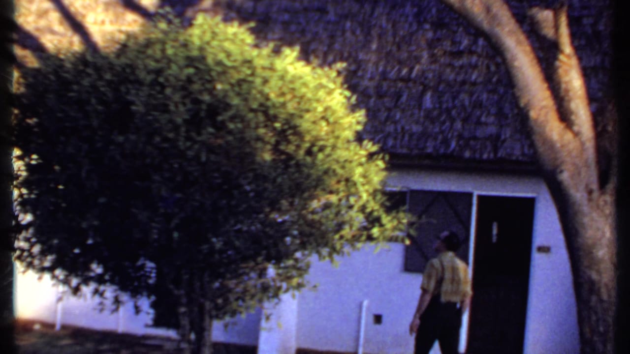 1969 Kenya home-movie frame showing a man crossing the exterior grounds near a dense shrub, tree, doorway, and thatched roof.