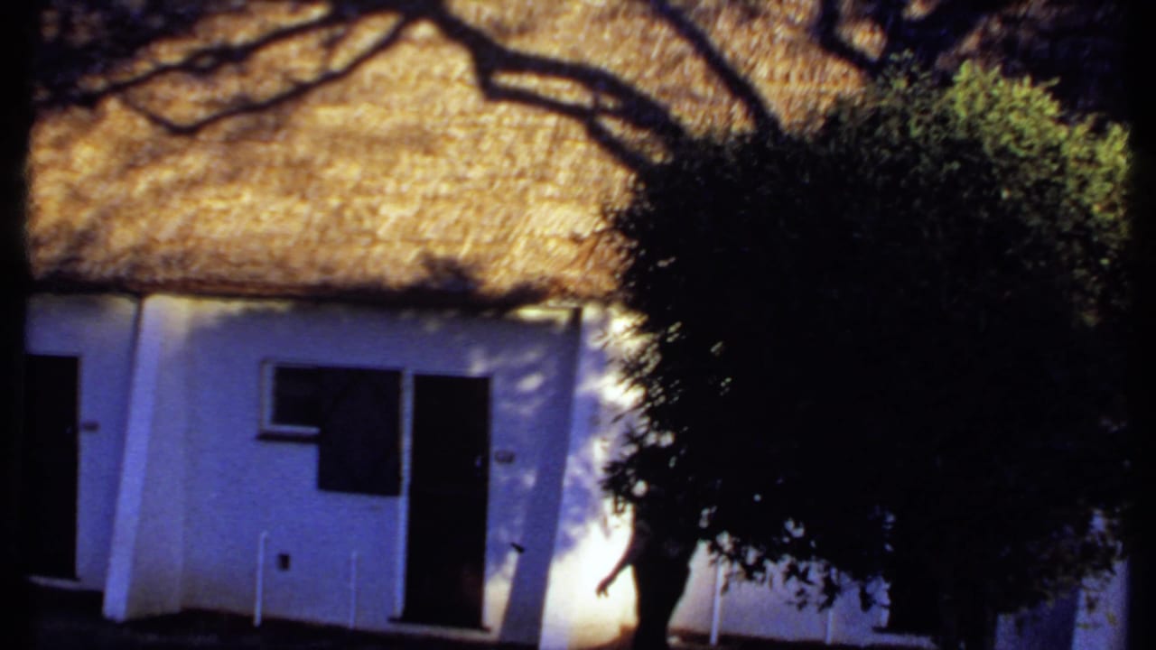 1969 Kenya home-movie frame with a white cottage, thatched roof, dark openings, and a large tree obscuring part of the facade.