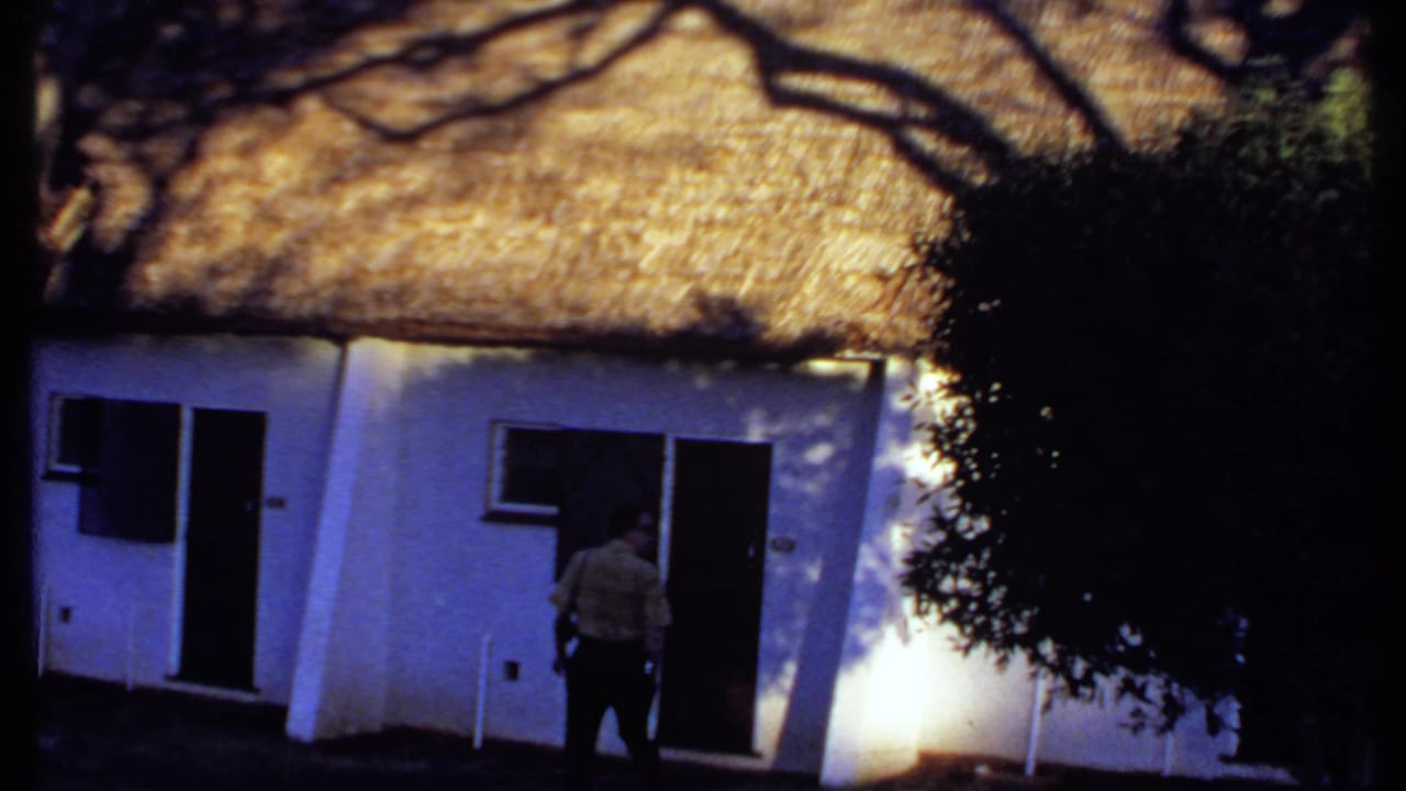 1969 Kenya 8mm frame showing a man near a white thatched cottage doorway, with tree shadows across the wall.