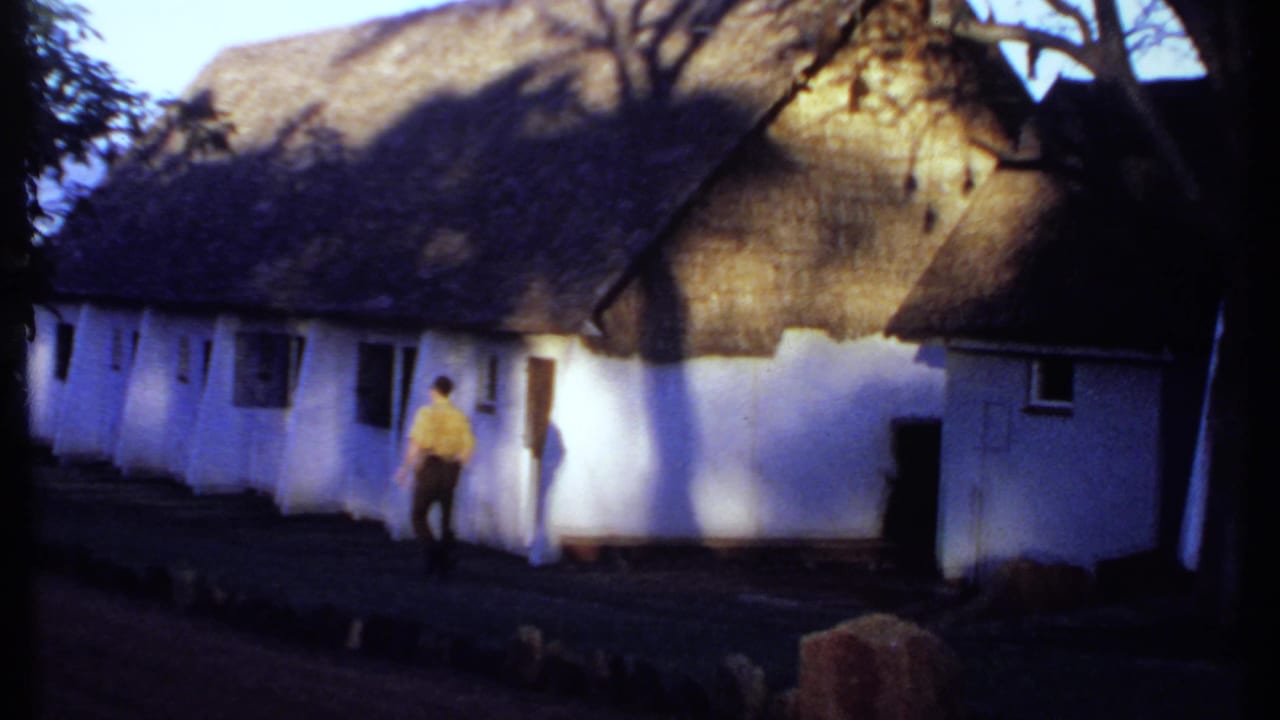 1969 Kenya frame showing a man in a yellow shirt walking beside white thatched cottages and a shaded path.