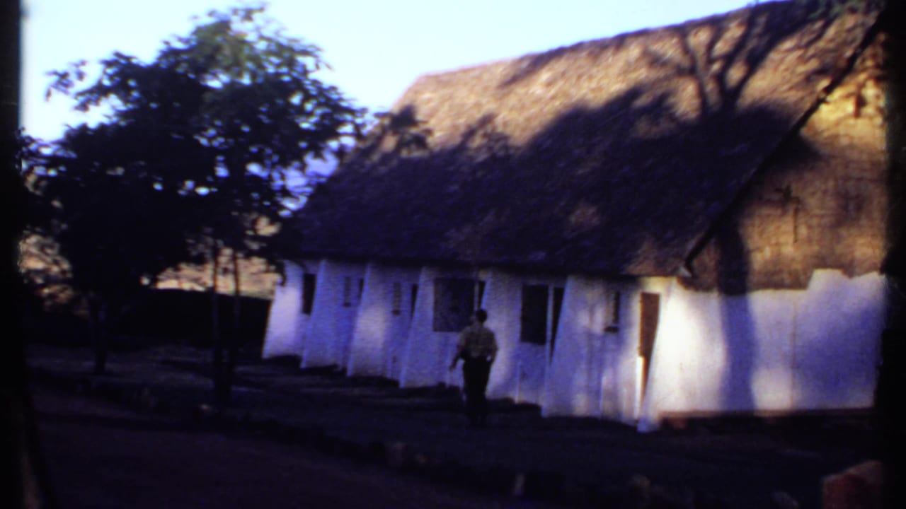 1969 Kenya home-movie frame with a man walking along a path beside a white thatched-roof structure and nearby trees.