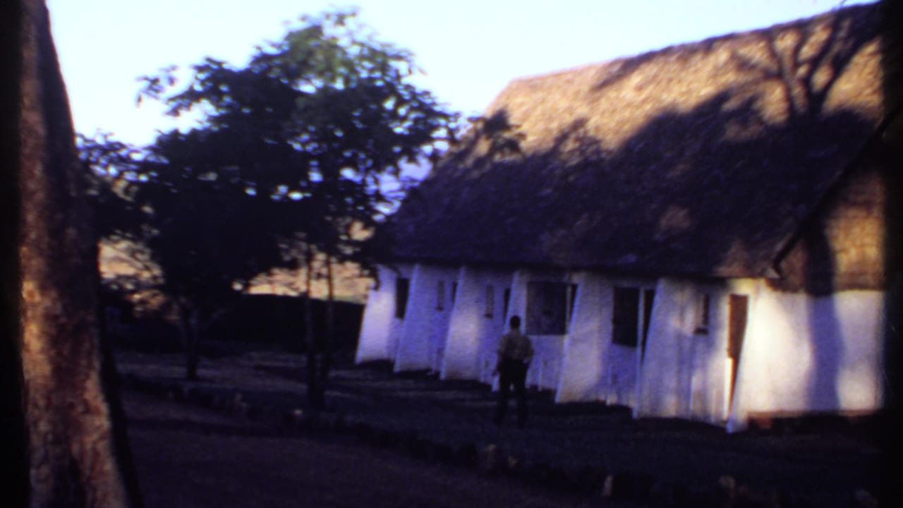 1969 Kenya 8mm frame showing a whitewashed thatched building framed by tree trunks as a man stands or walks near the facade.