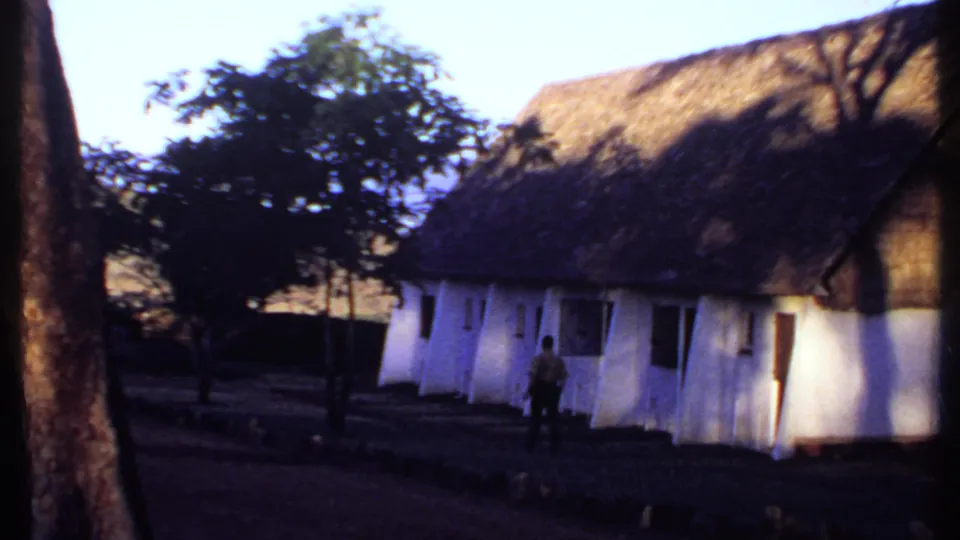 1969 Kenya 8mm frame showing a whitewashed thatched building framed by tree trunks as a man stands or walks near the facade.