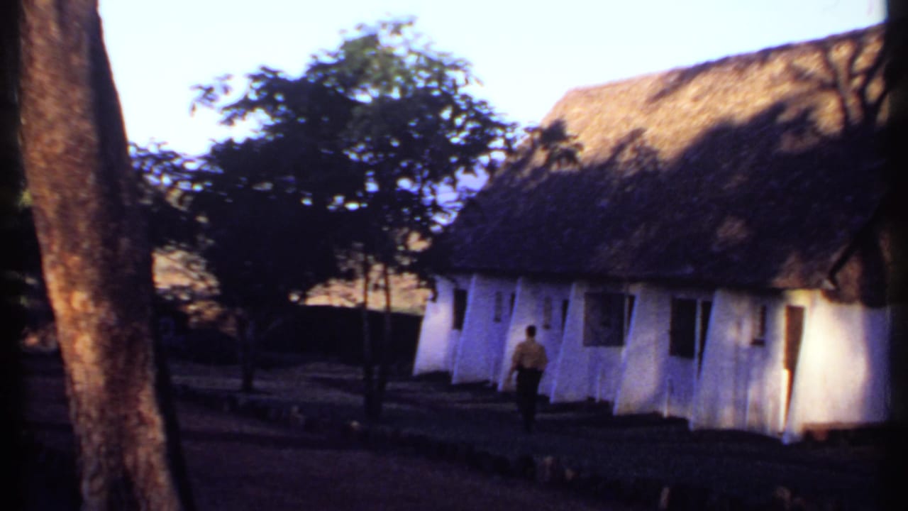 1969 Kenya frame of a man in a yellow shirt passing the shaded side of a white building with a thick thatched roof.