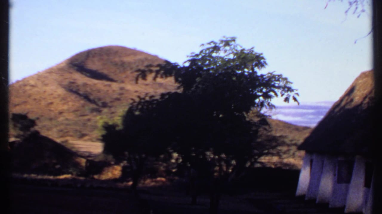 Dry rounded hill behind silhouetted trees and thatched structures.