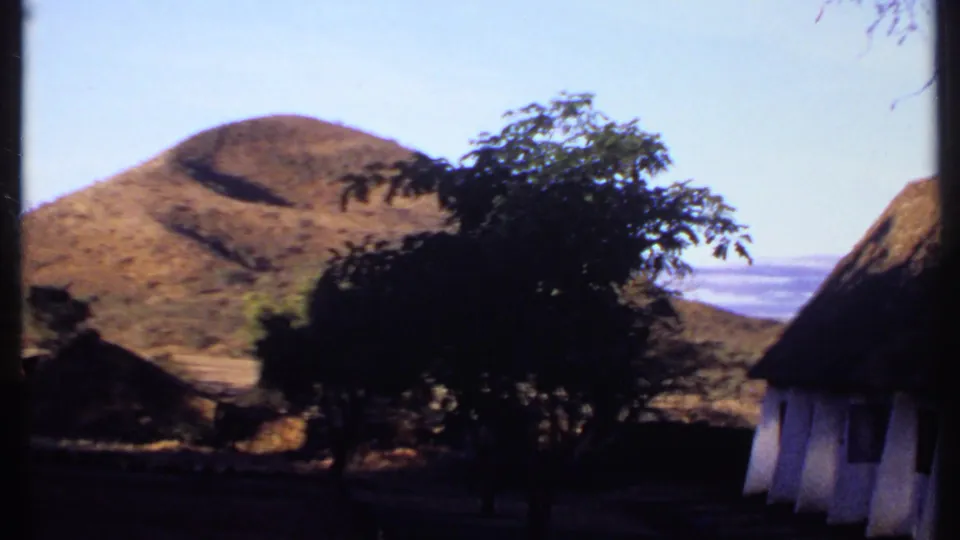 1969 Kenya 8mm frame showing a dry rounded hill, dark foreground trees, and thatched roofs under pale sky.