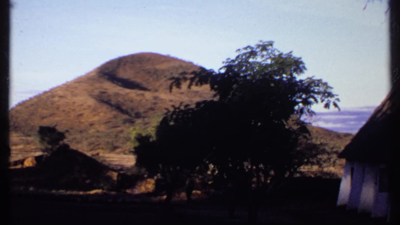 1969 Kenya 8mm frame with a sunlit dry hill partly framed by silhouetted trees and a thatched roof edge.