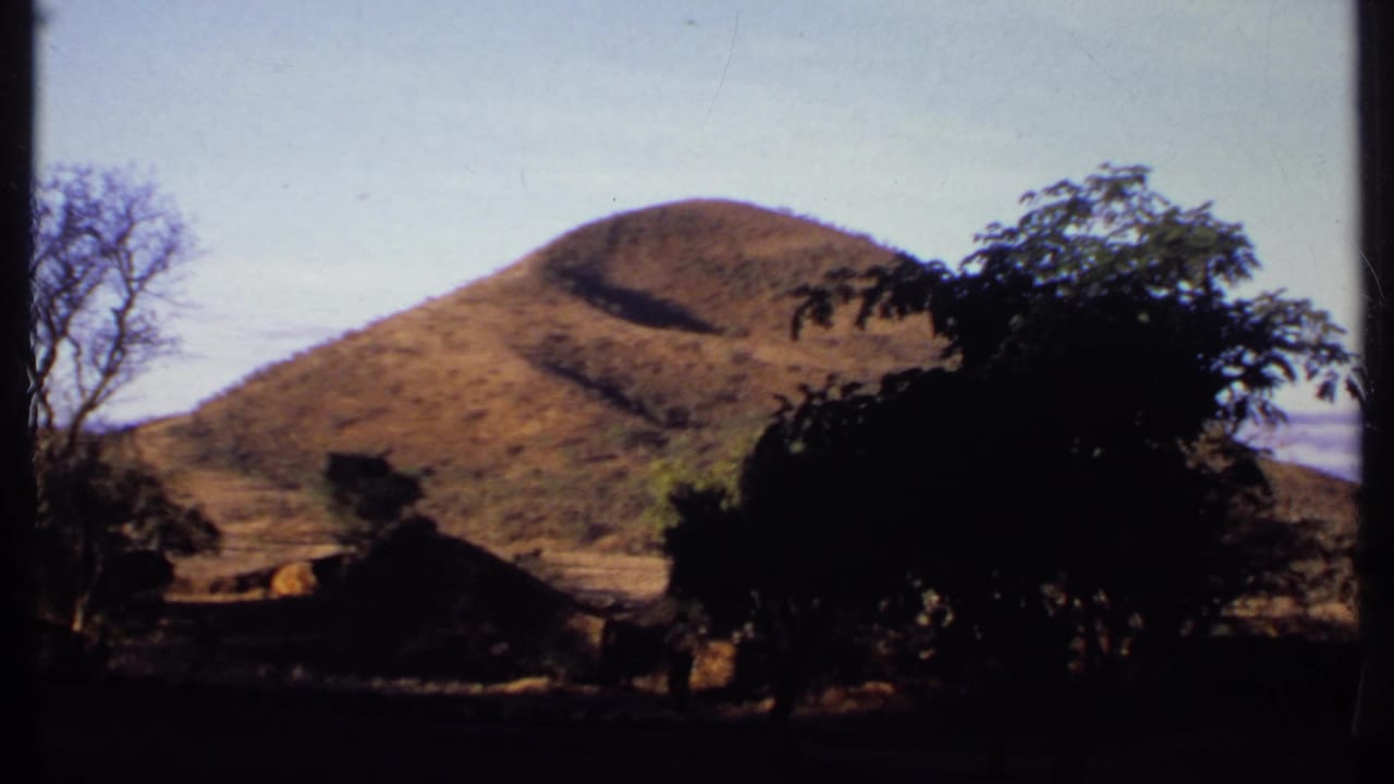 1969 Kenya 8mm frame with a dry hill beyond trees, thatched roofs, and a small white-walled structure edge.