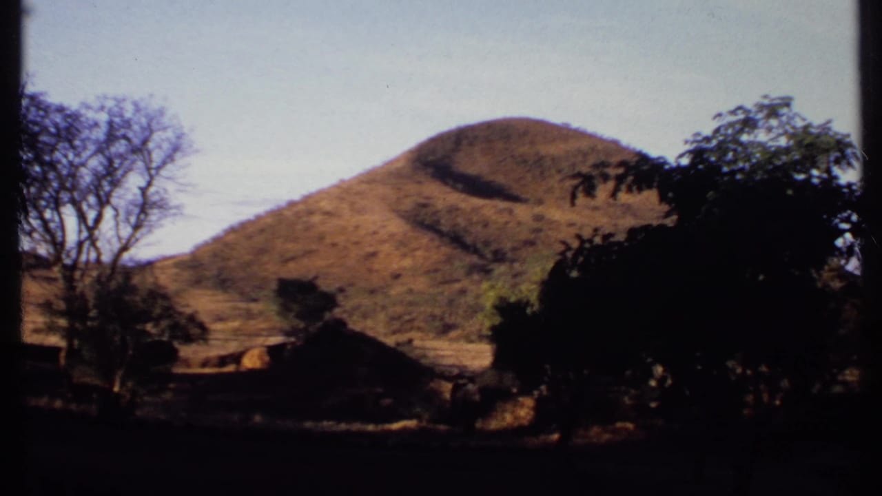 1969 Kenya home-movie frame showing a sunlit ochre hill, dark tree silhouettes, a shaded foreground, and low structures at the base.