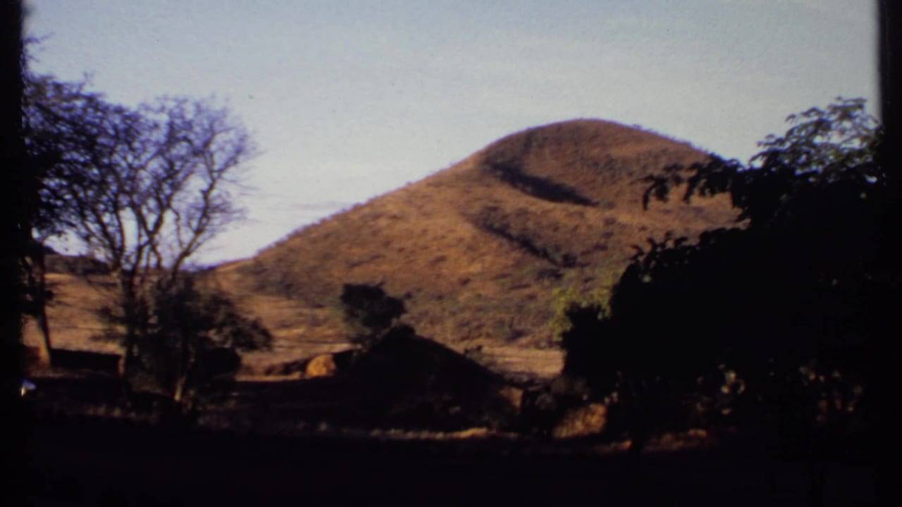 1969 Kenya 8mm frame of a warm-toned treeless hill seen beyond sparse trees, deep shade, and faint structures near the slope.