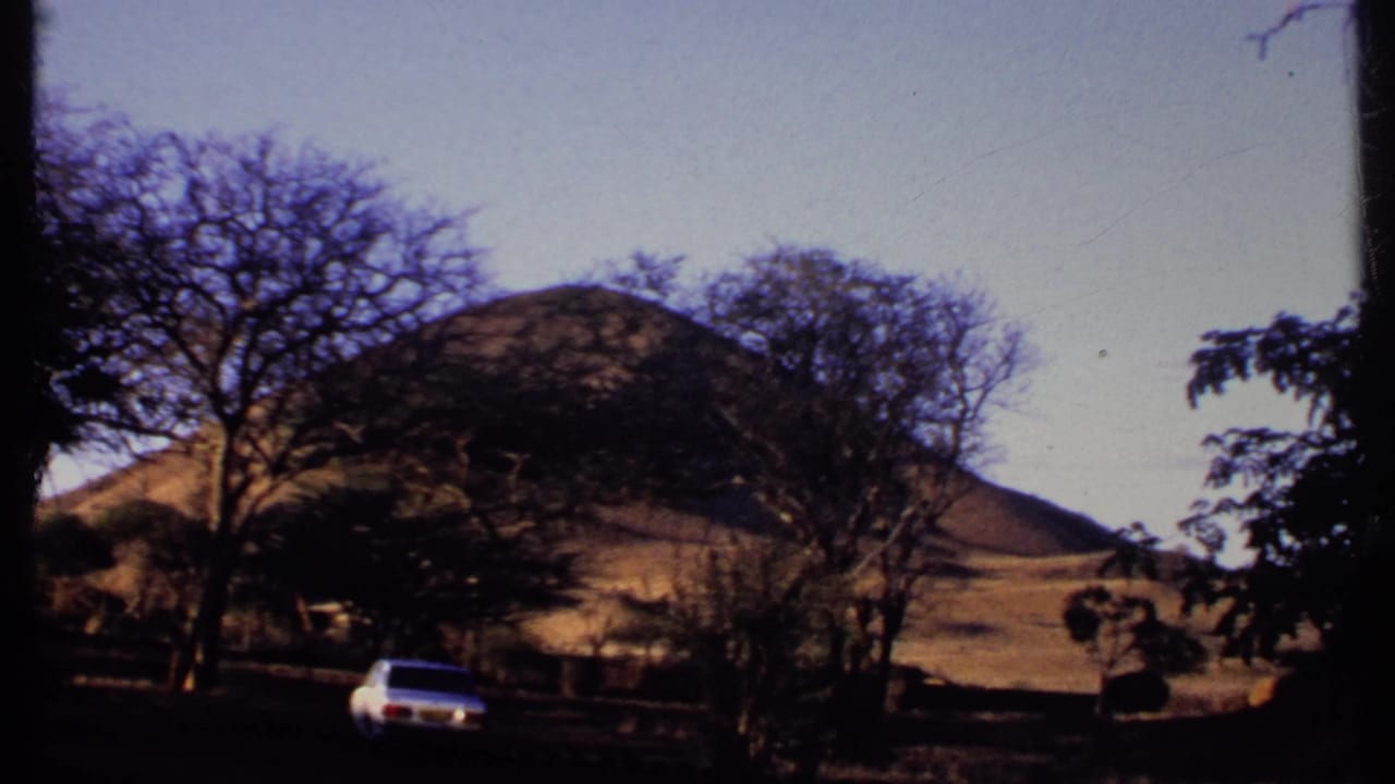 1969 Kenya 8mm view of a white sedan on a rural road, framed by trees and dry hills.