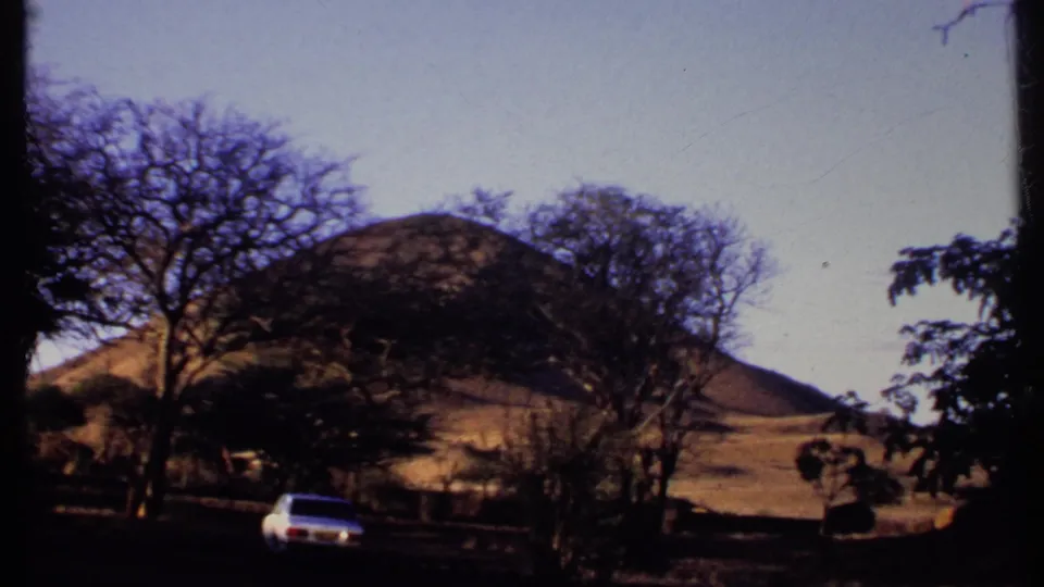 1969 Kenya 8mm view of a white sedan on a rural road, framed by trees and dry hills.