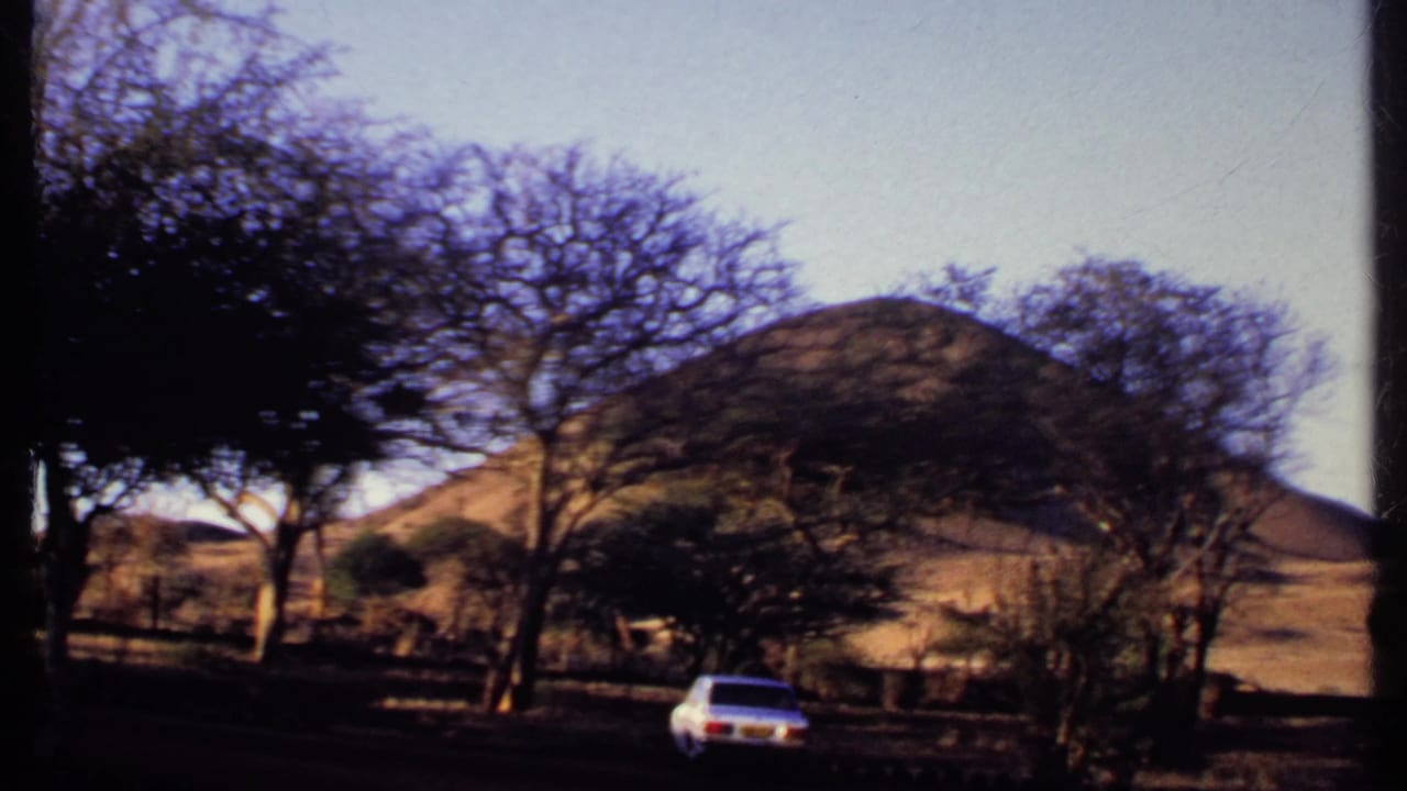1969 Kenya home-movie frame of a white sedan crossing a shaded road below dry hills and trees.