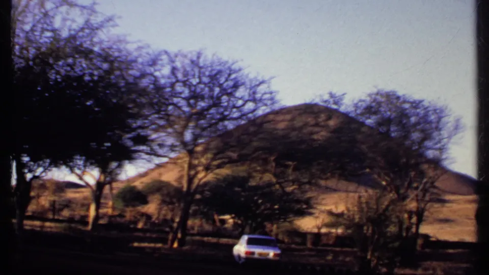 1969 Kenya home-movie frame of a white sedan crossing a shaded road below dry hills and trees.