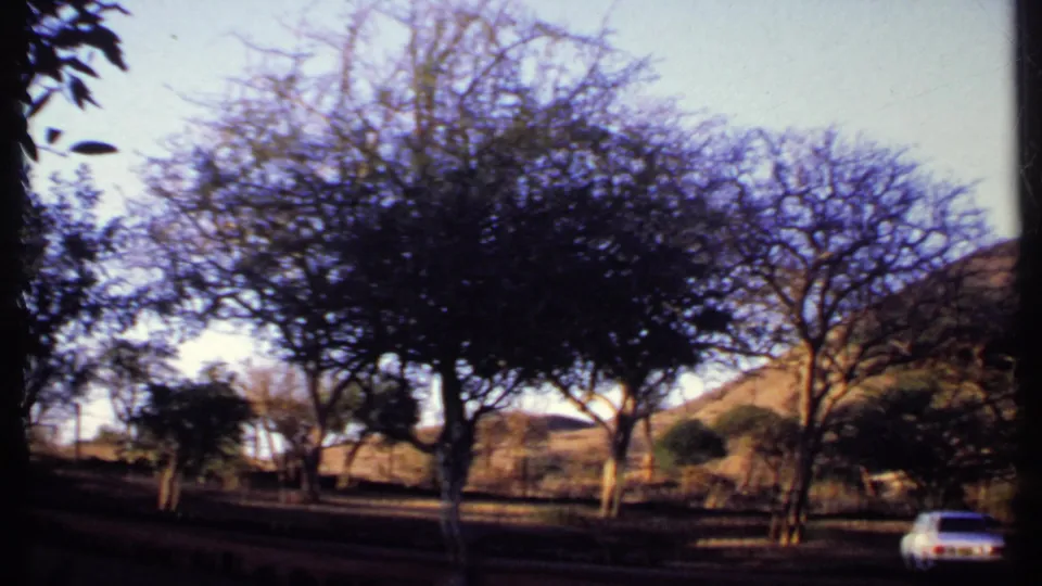 A 1969 Kenya roadside view from moving 8mm footage, with a white car, sparse trees, and rolling dry hills.