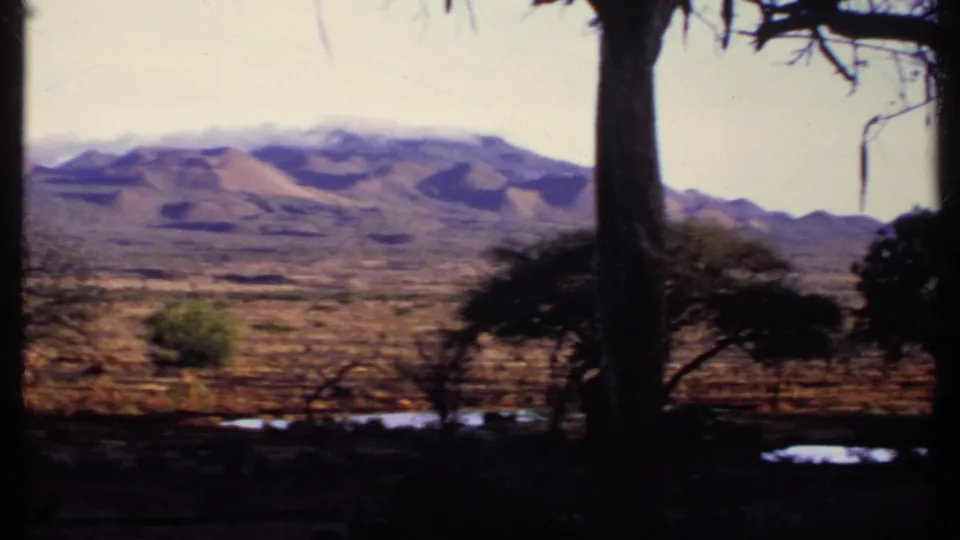 1969 Kenya 8mm view of distant mountains, dry open terrain, roadside tree trunks, and small reflective water patches.