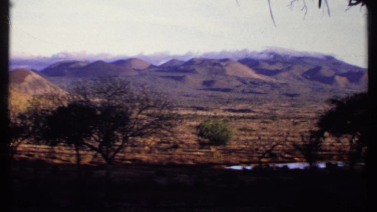 1969 Kenya landscape frame with scrubby foreground trees, reflective water patches, and a layered mountain range across the horizon.