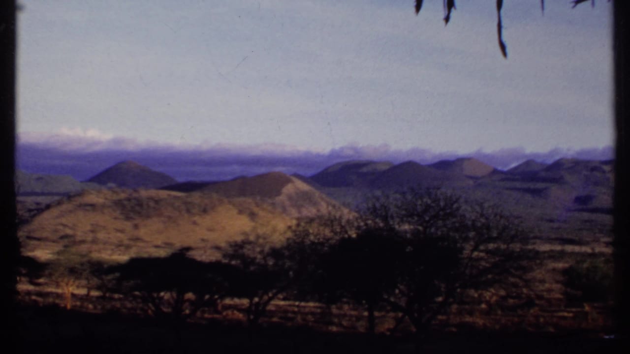 1969 Kenya home-movie frame with dark foreground shrubs, arid terrain, rounded hills, distant ridges, and a pale open sky.