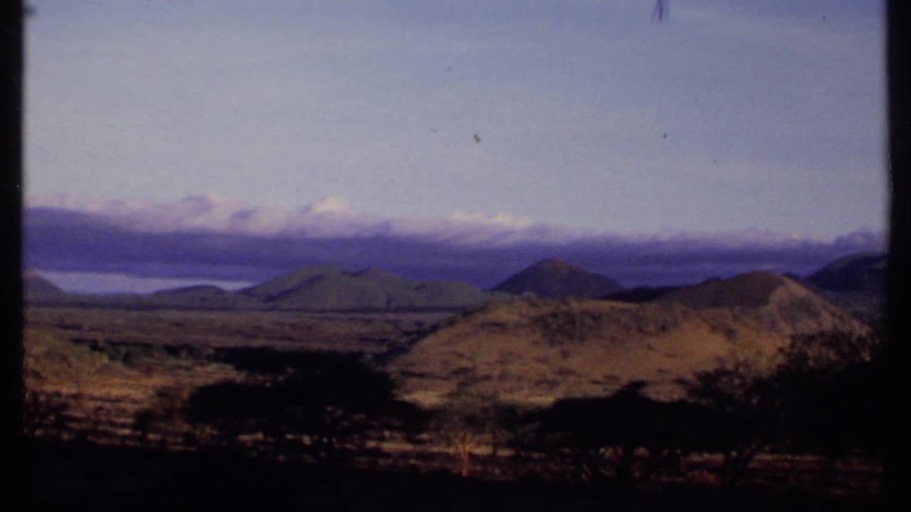 1969 Kenya 8mm frame looking over arid foreground and acacia-like silhouettes toward hills, possible water, and low clouds.