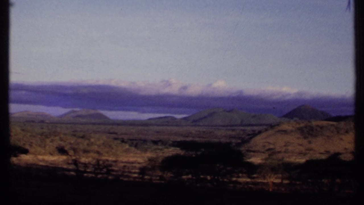 1969 Kenya 8mm frame showing rounded hills, dark foreground trees, a possible distant lake, and cloud-covered ridges.