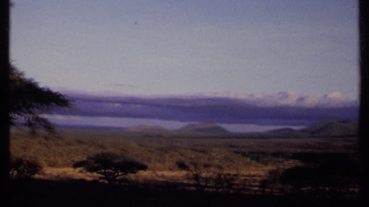 1969 Kenya 8mm frame with foreground scrub and acacia-like trees overlooking a pale distant water body, hills, and clouds.