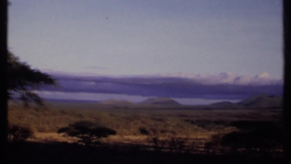 1969 Kenya 8mm frame with foreground scrub and acacia-like trees overlooking a pale distant water body, hills, and clouds.