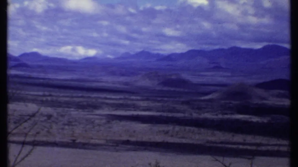 1969 Kenya travel footage frame with a broad dry valley, layered mountain ridges, and visible 8mm frame bars.