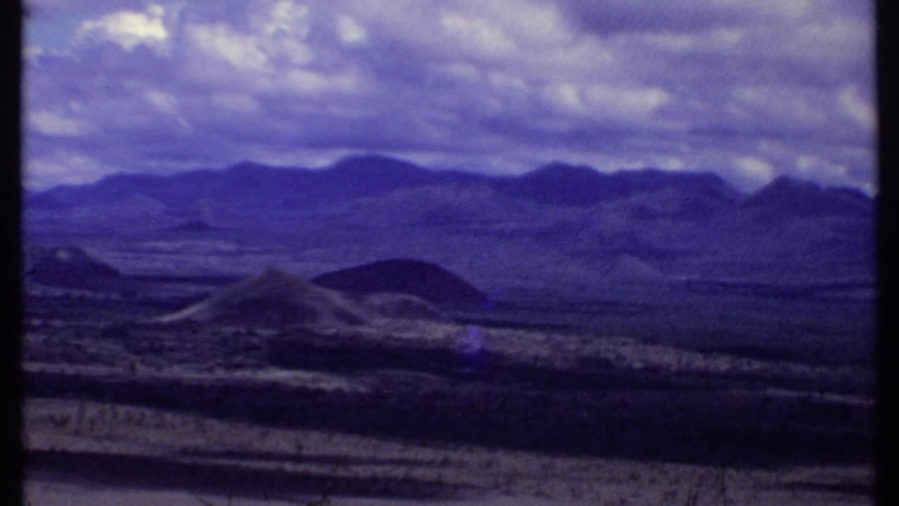 1969 Kenya 8mm frame showing dry open ground, low dark hills, distant mountains, and a cloudy sky during a slow pan.