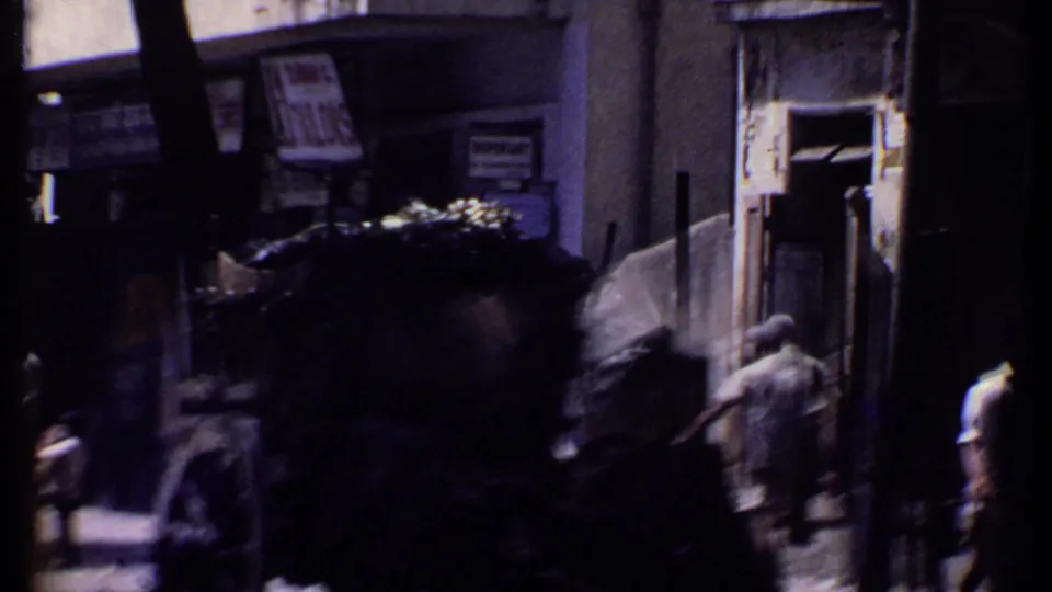 1969 Kenya 8mm frame of a shaded street with pedestrians near a covered cart and storefronts, with signage visible but not clearly readable.