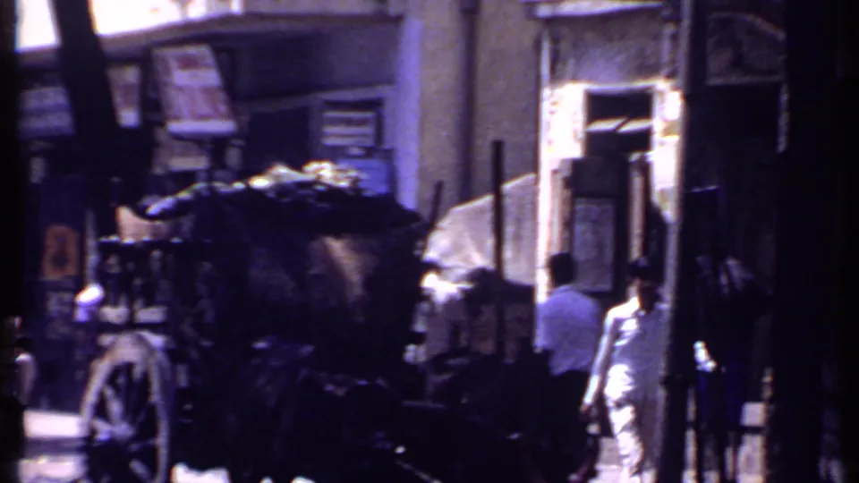 1969 Kenya 8mm frame with pedestrians walking beside a covered cart and building facades, filmed in high contrast shade.