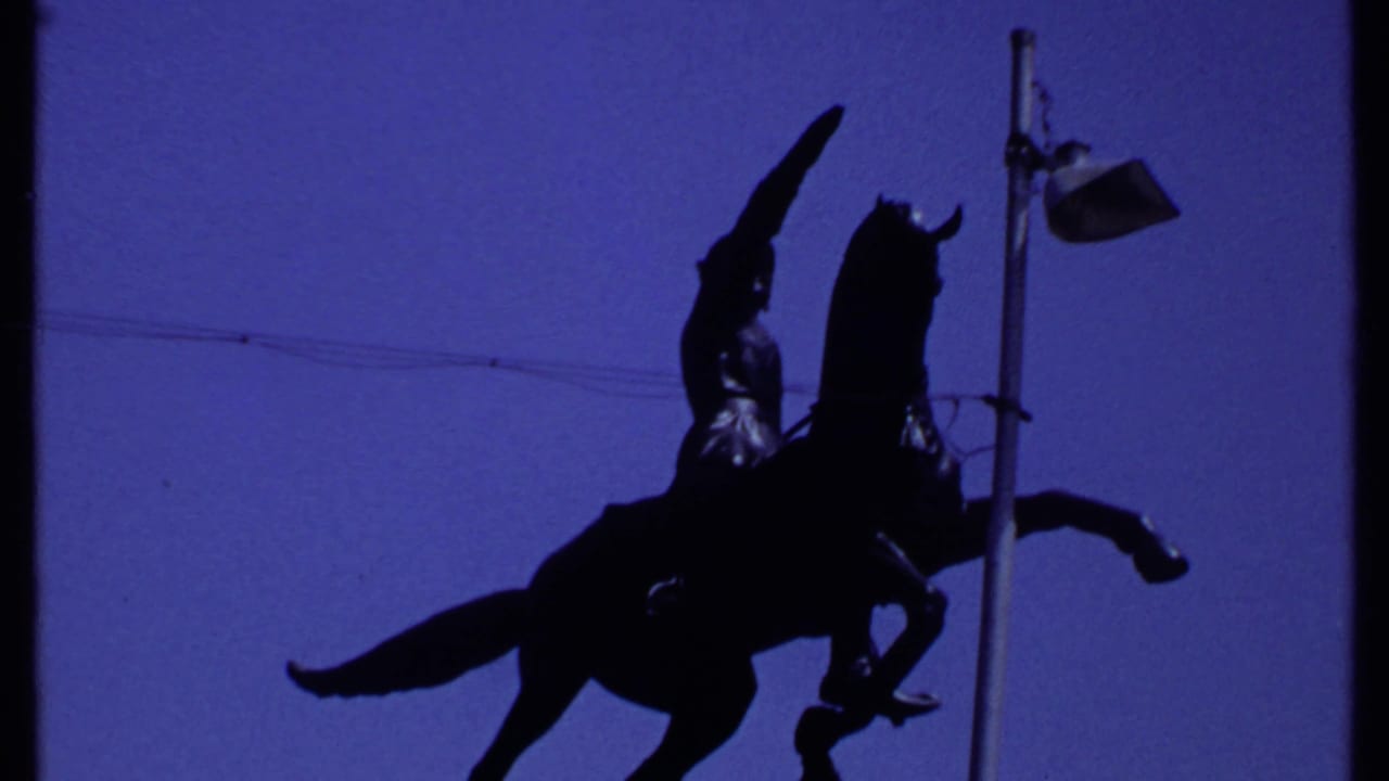 1969 Kenya 8mm frame showing a dark mounted warrior statue silhouetted against blue sky beside a streetlamp.
