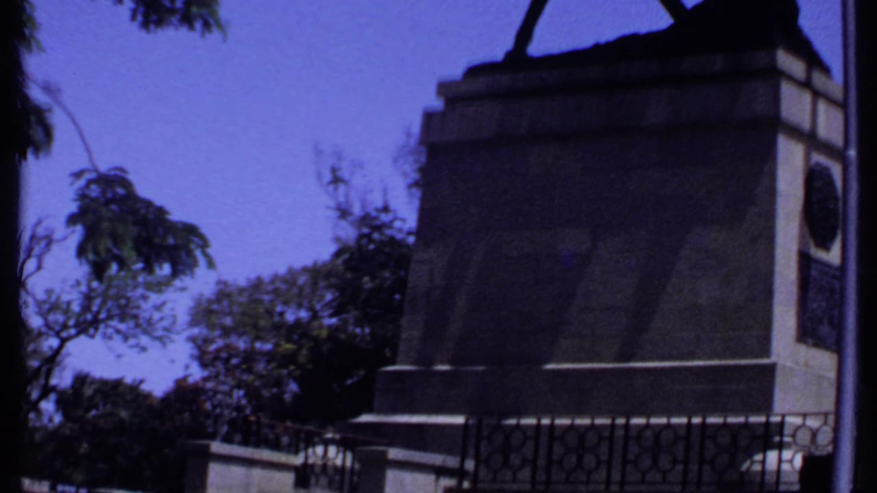 1969 Kenya 8mm frame showing a stone monument base, iron railings, trees, and a clear blue sky in a public outdoor setting.