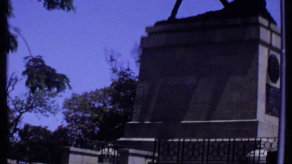 1969 Kenya 8mm frame showing a stone monument base, iron railings, trees, and a clear blue sky in a public outdoor setting.