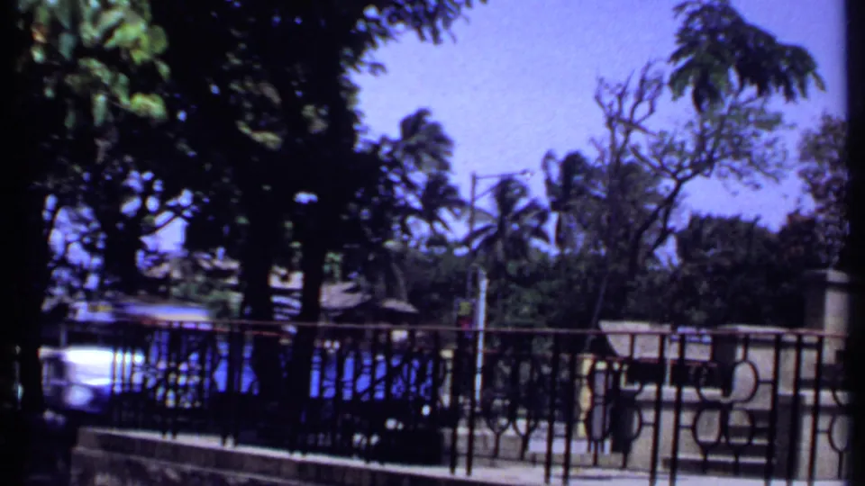 1969 Kenya 8mm frame with a blue truck moving along a road behind iron railings, trees, and stone gate structures.