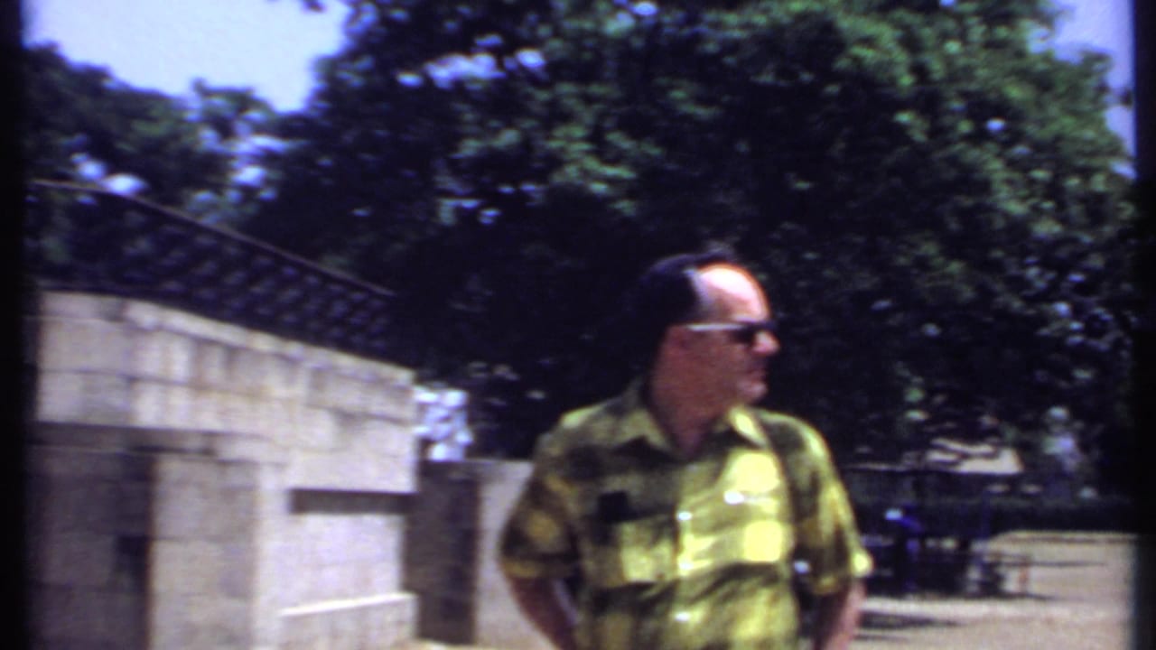 Man in sunglasses standing near stone monument walls and trees.