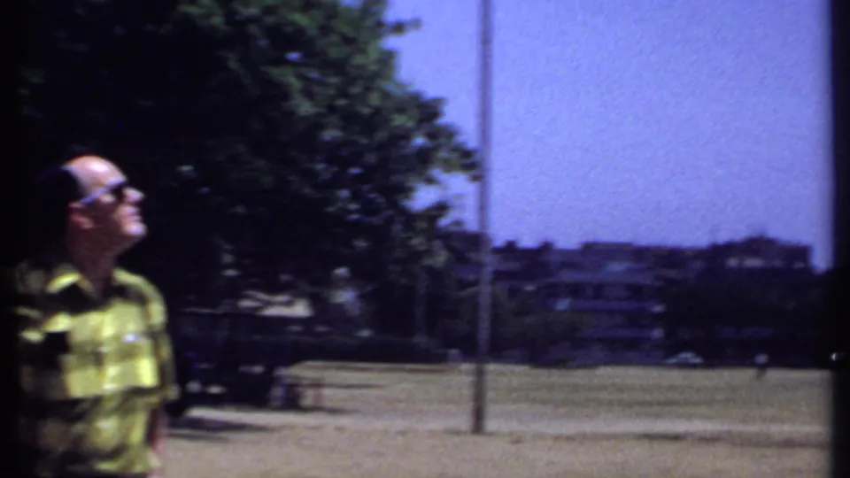 1969 Kenya 8mm frame showing a man in sunglasses near a large open field with trees and distant buildings.