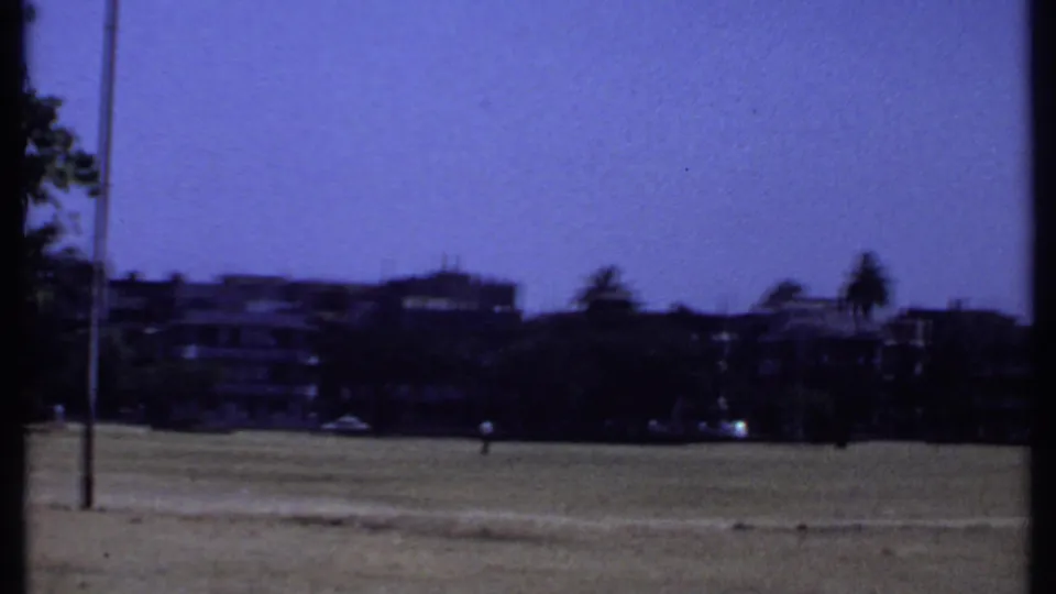 1969 Kenya home-movie frame of an open field, distant low buildings, trees and blue sky.