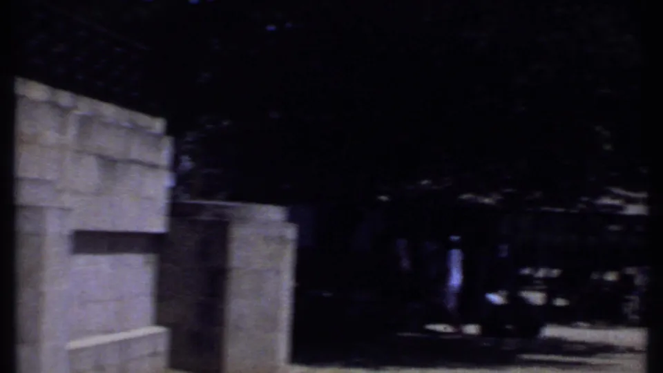 1969 Kenya 8mm frame showing a shaded park edge with a pale wall, trees, posts, picnic tables, and distant people.