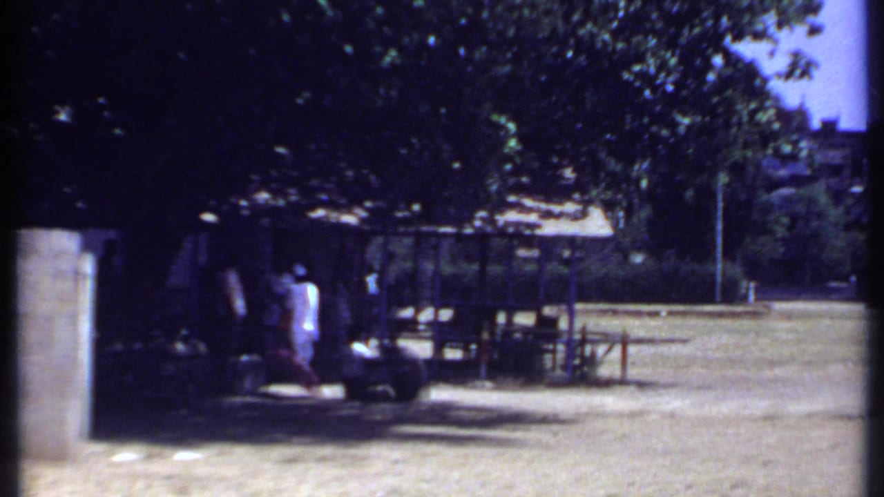 People near a shaded shelter with tables, trees, open ground, and a parked motorcycle-like vehicle.