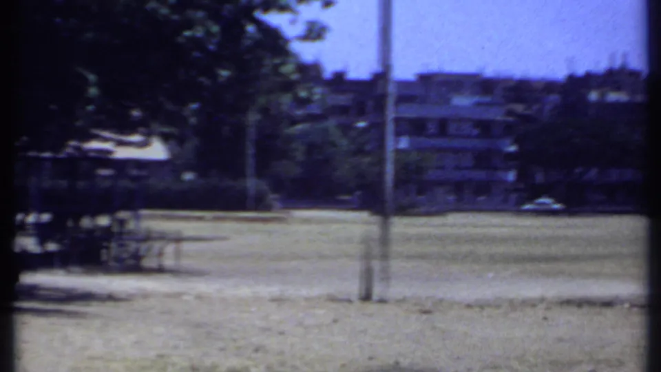 1969 Kenya 8mm frame looking across an open dry field toward shade trees, posts, picnic tables, and distant low-rise buildings.