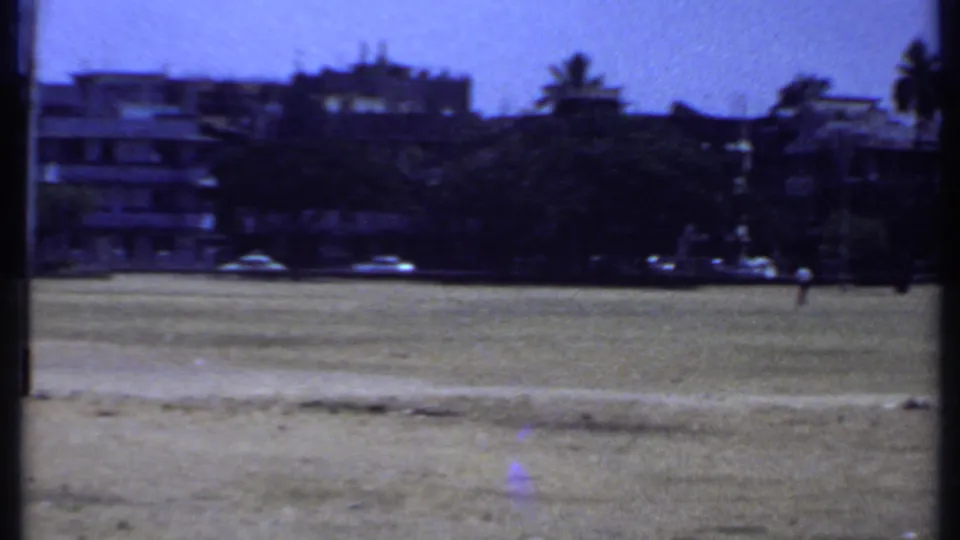 1969 Kenya 8mm frame showing a wide open field, background buildings, parked cars, trees, palm silhouettes, and a distant person.