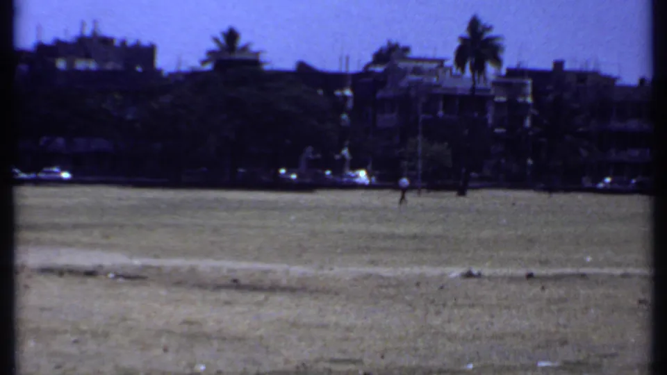 1969 Kenya home-movie frame showing open ground, distant low-rise buildings, palm trees, and small figures near the horizon.