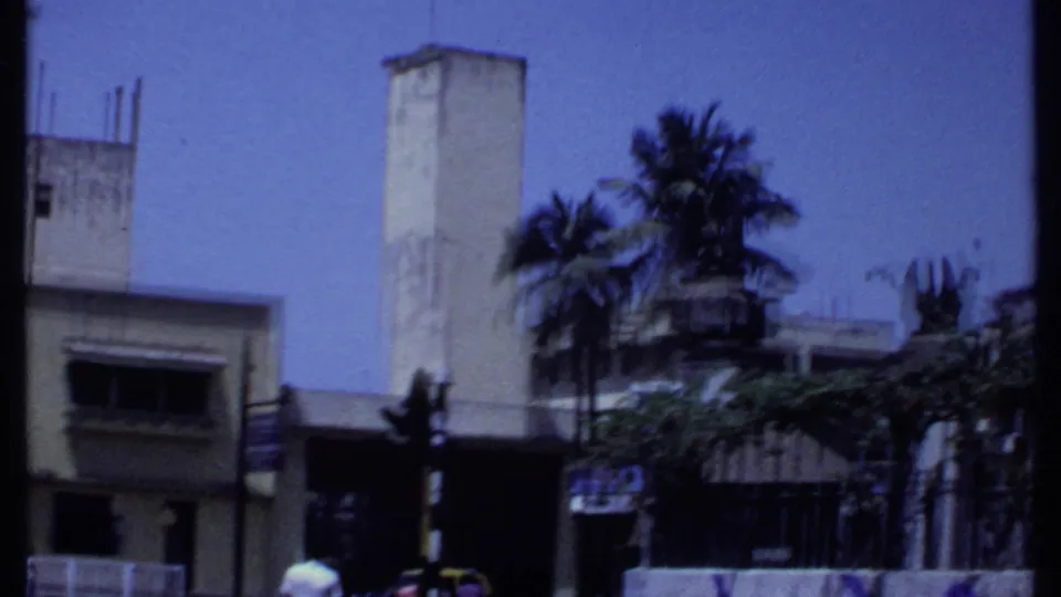 1969 Kenya street view with a pale tower-like building, palms, roadside structures, and small people near the lower edge.