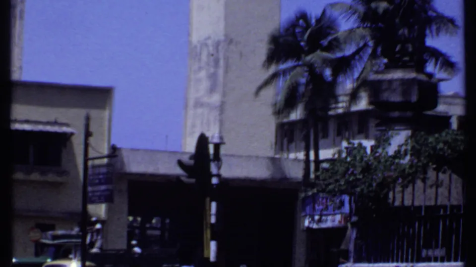 1969 Kenya home-movie view of a street corner with a concrete building, palm trees, fencing, and partially readable signs.