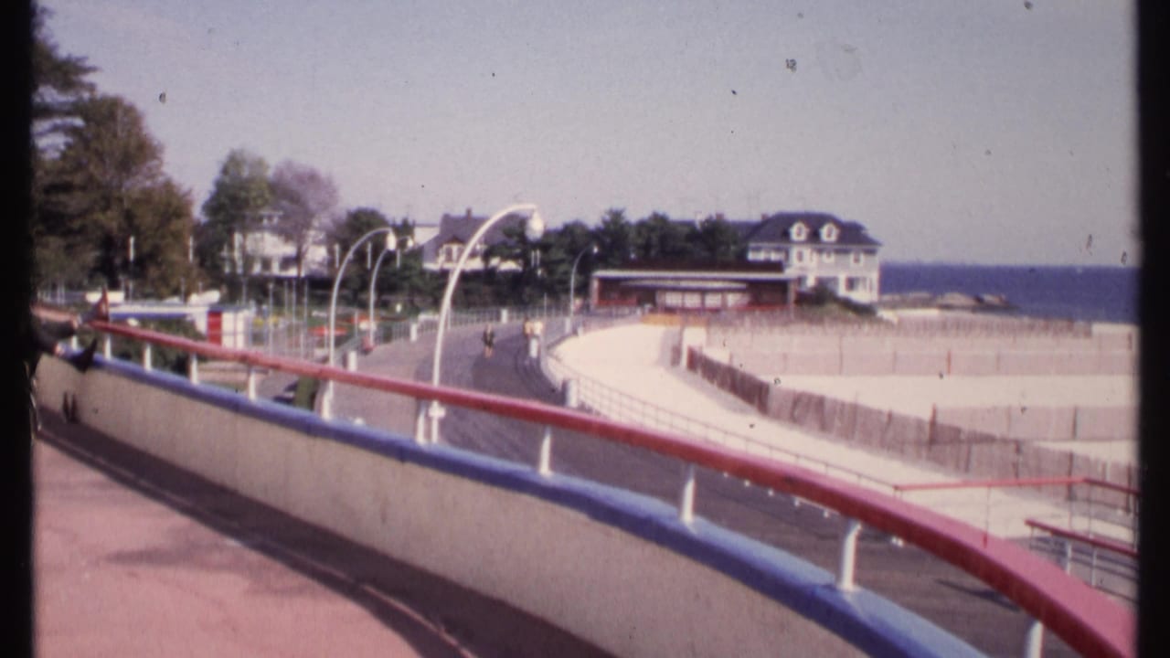 1969 New London shoreline view from a curved boardwalk with red and blue railings, beach fencing, small pedestrians, houses, and ocean beyond.