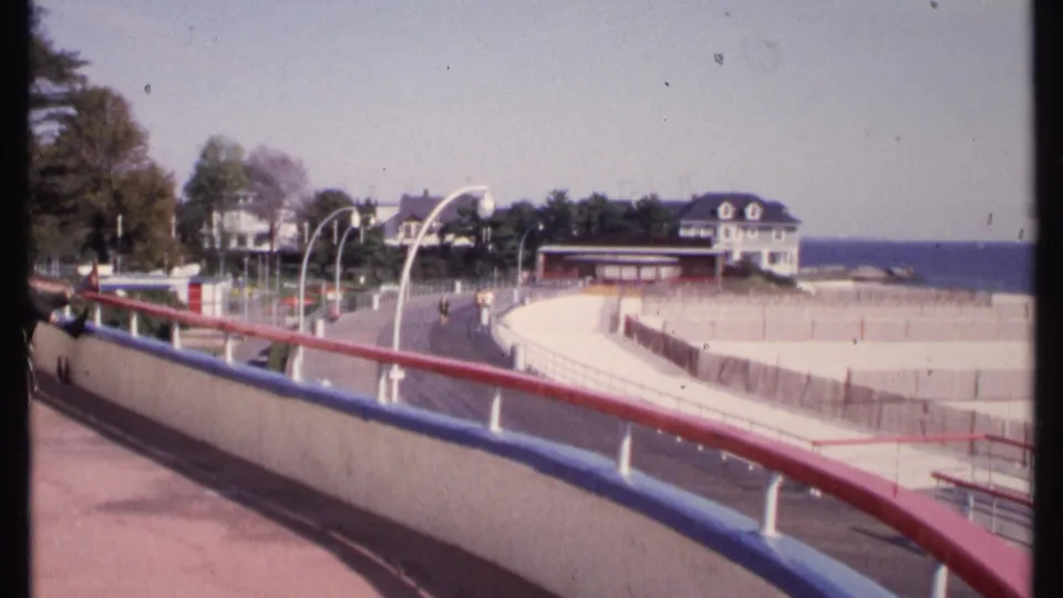 1969 New London shoreline view from a curved boardwalk with red and blue railings, beach fencing, small pedestrians, houses, and ocean beyond.