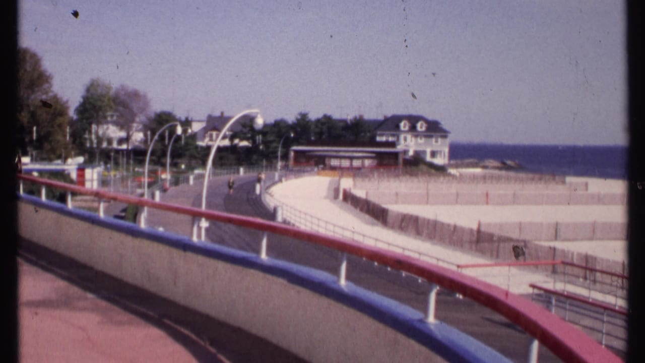 Boardwalk and railings frame a beach area, road, white houses, trees, and ocean horizon.